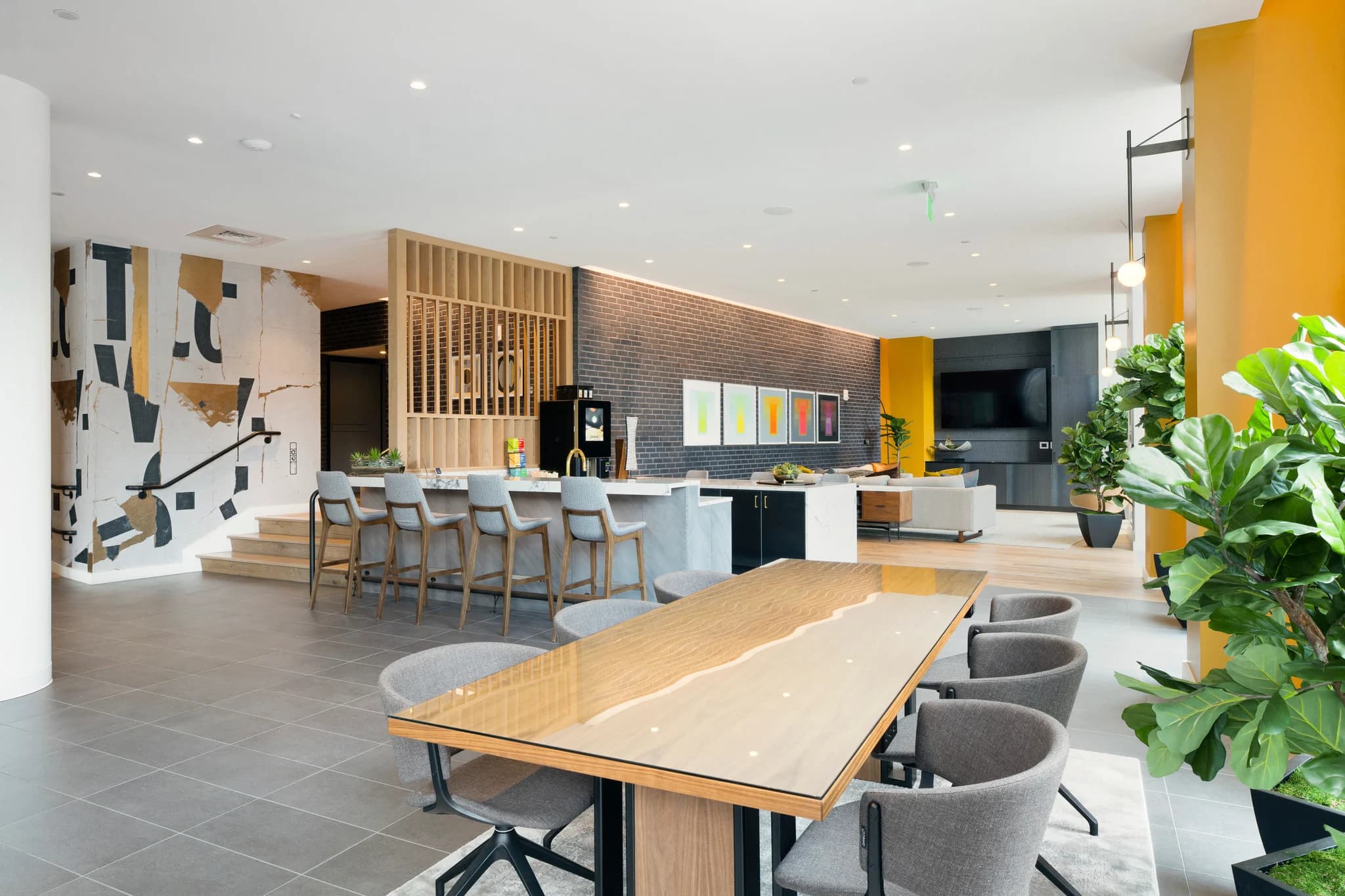 Open-concept common room with long wooden dining table, bar counter with seating, gray upholstered chairs, yellow accent walls, and potted plants.