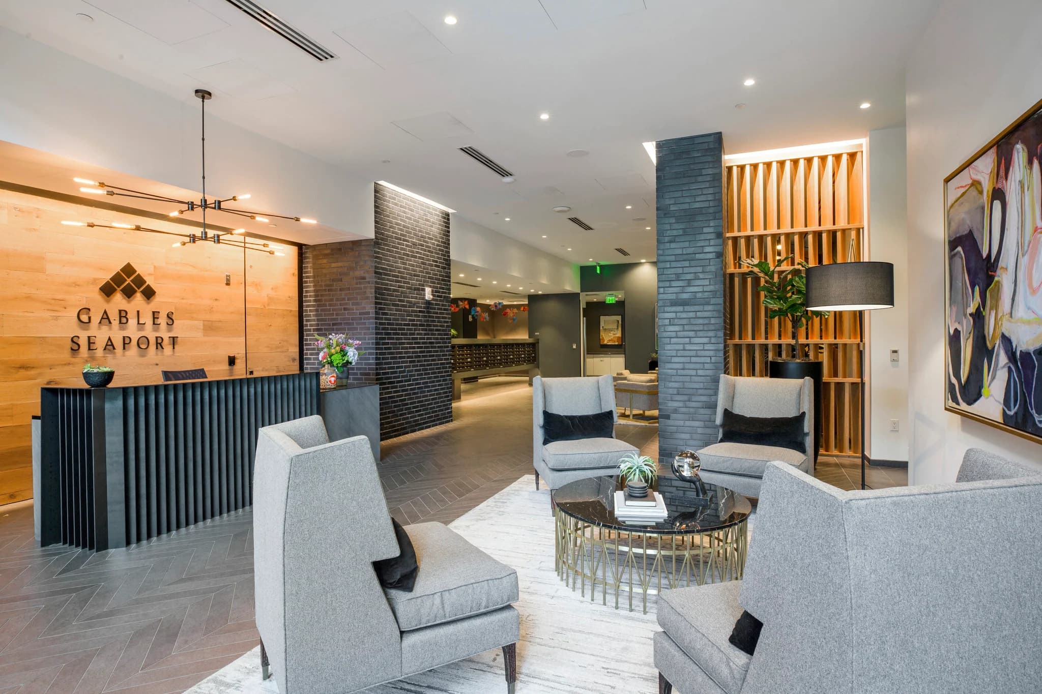Modern apartment lobby with gray upholstered chairs, round black table, wood-paneled walls, and reception desk with Gables Seaport signage.