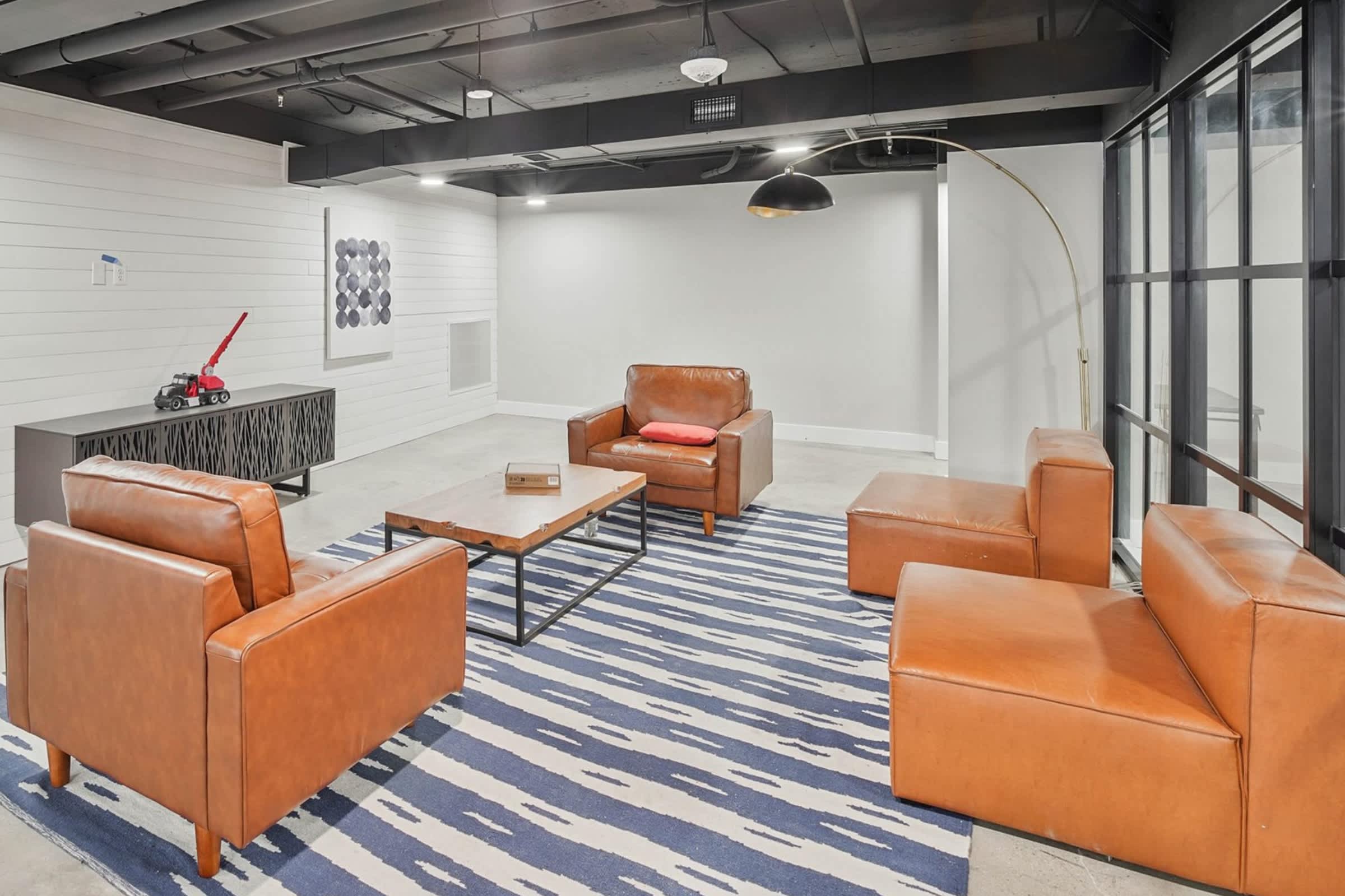 Common lounge with four cognac leather chairs arranged around a white coffee table, blue and white striped rug, arc floor lamp, and large windows.