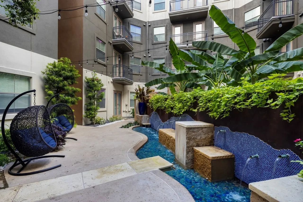 Courtyard with hanging egg chair, tropical plants, banana trees, blue water feature, stone seating, and multi-story residential building.