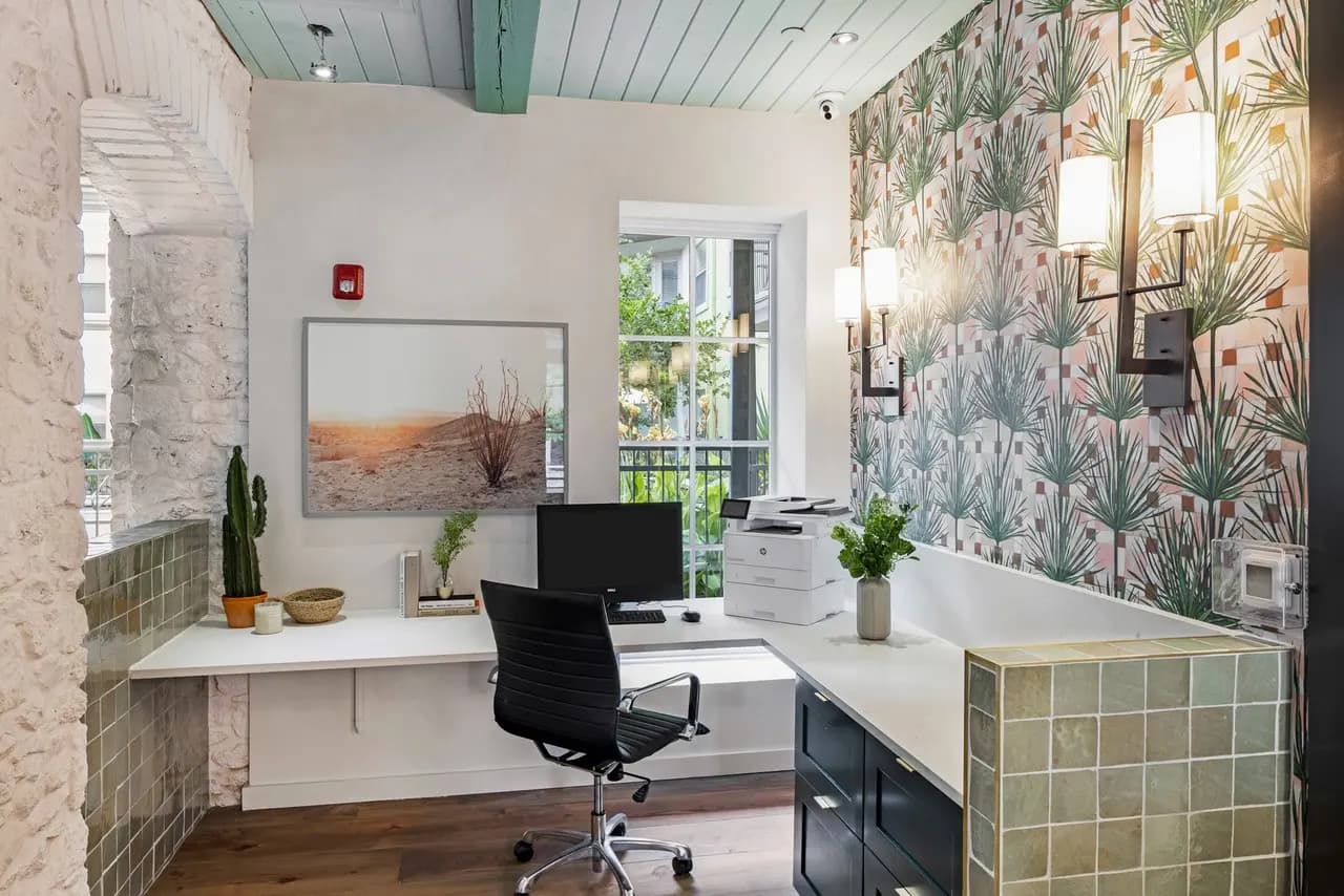 Home office with L-shaped white desk, black office chair, botanical wallpaper accent wall, and glass doors opening to a balcony.