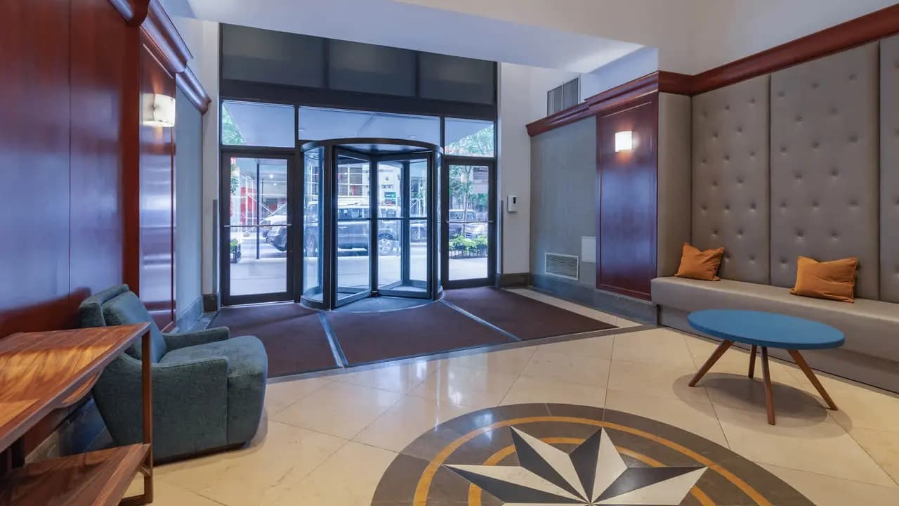Apartment building lobby with black-framed glass doors, beige tile flooring with geometric star pattern, gray upholstered seating, and wood accents.