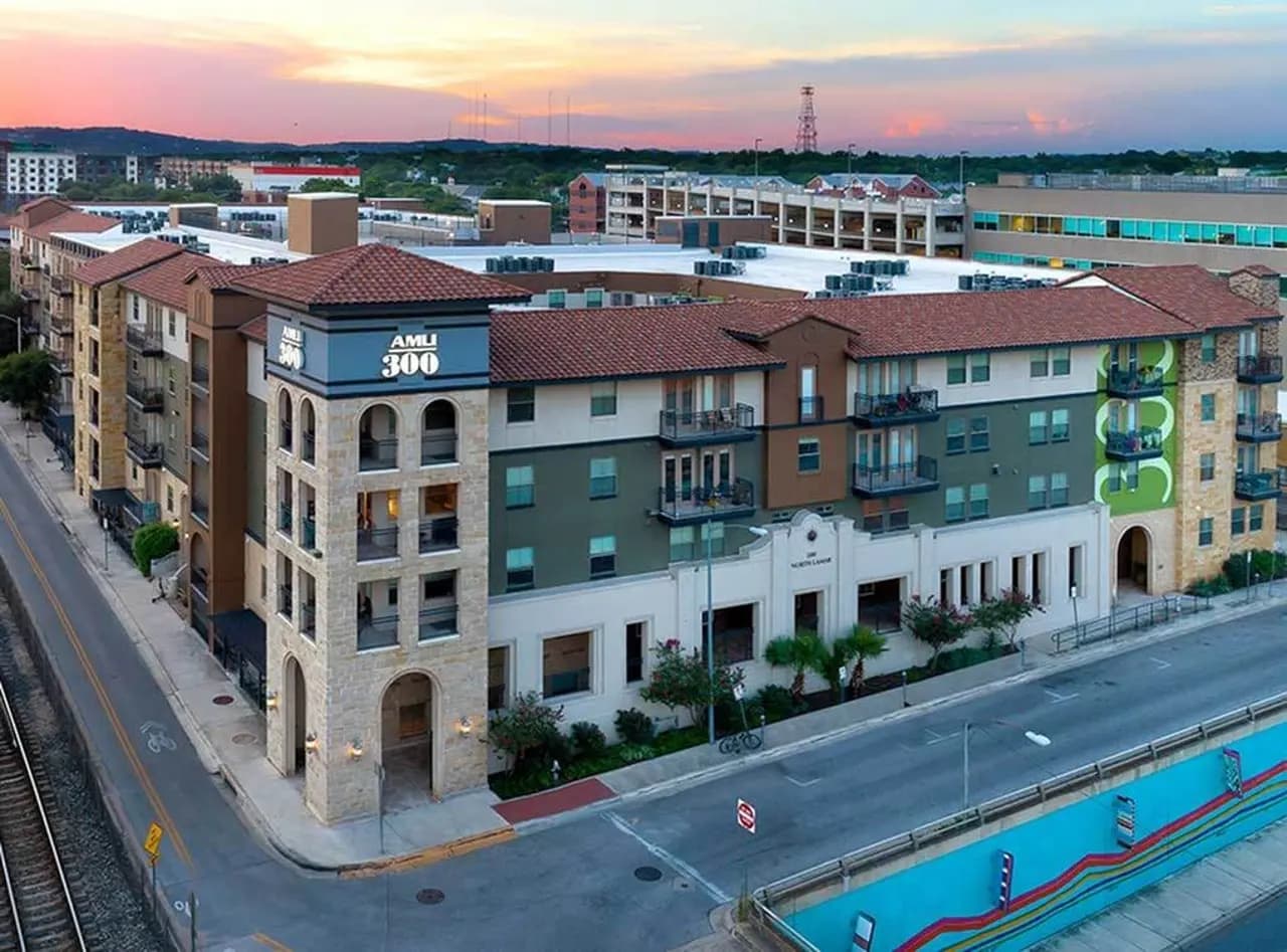 Multi-story residential building with brown and green facade, white ground-floor retail space, and urban surroundings at dusk.