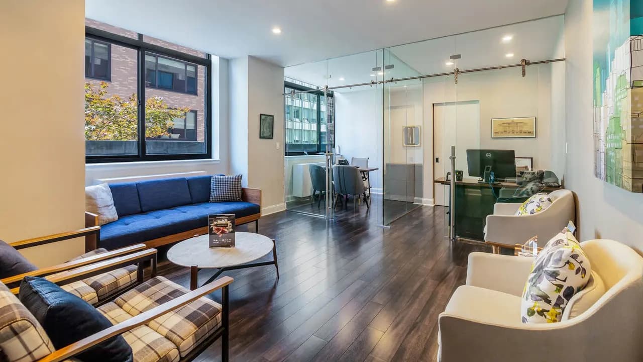 Open-plan living room with blue and plaid mid-century sofas, white round coffee table, dark wood flooring, large windows with city views, and glass partition walls.