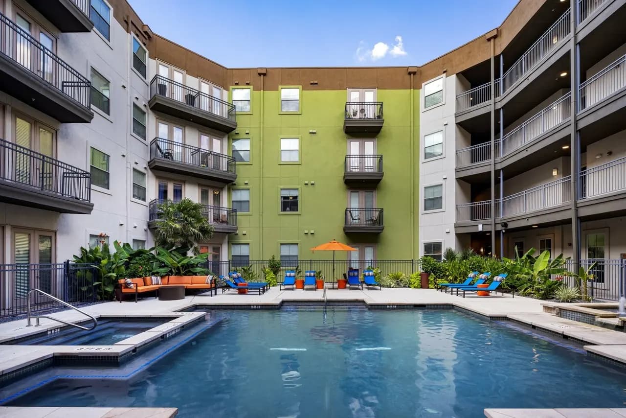 Courtyard pool with blue water, orange lounge chairs, tropical plants, and lime-green residential building with balconies under clear sky.