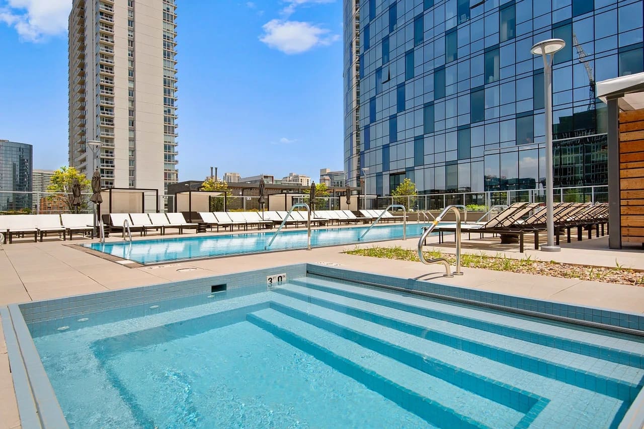 Rooftop pool area with lap pool in foreground, larger swimming pool, lounge chairs, and modern high-rise buildings against blue sky.