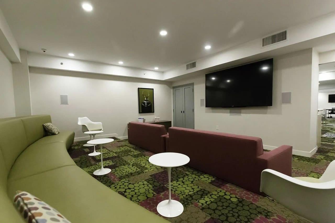 Modern lounge with green and burgundy sofas, white accent chairs, patterned carpet, wall-mounted TV, and recessed lighting.