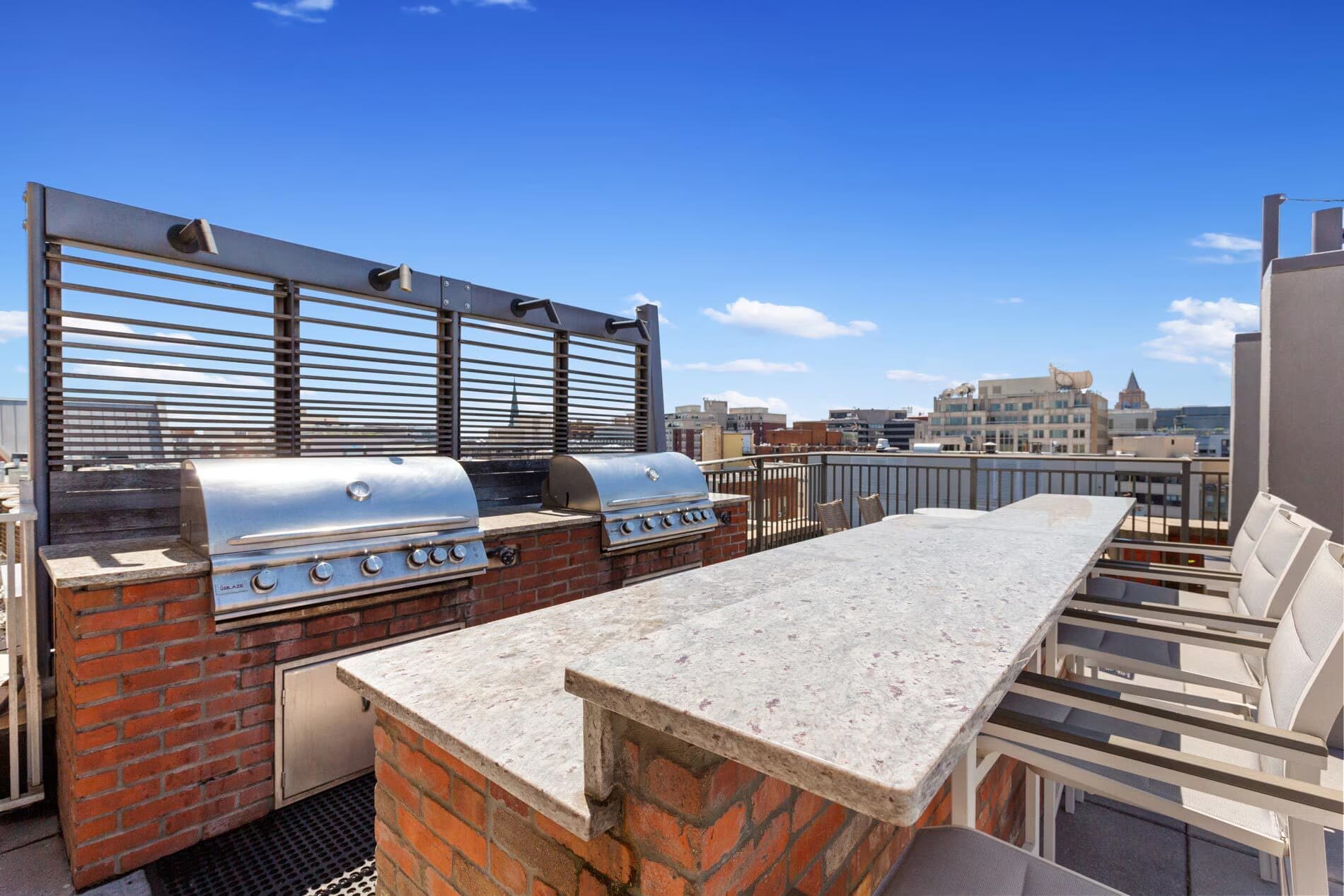 Rooftop terrace with stainless steel grills, brick counter, lounge chairs, and downtown Washington DC skyline view under clear blue sky.
