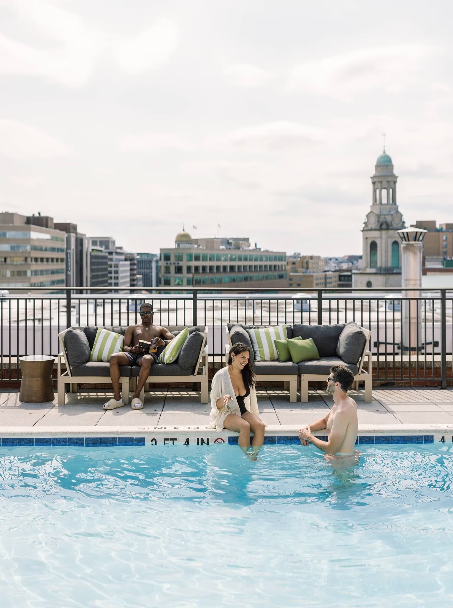 Rooftop pool with two people in water, lounge seating with green and gray pillows, and Washington DC skyline with historic dome building in background.