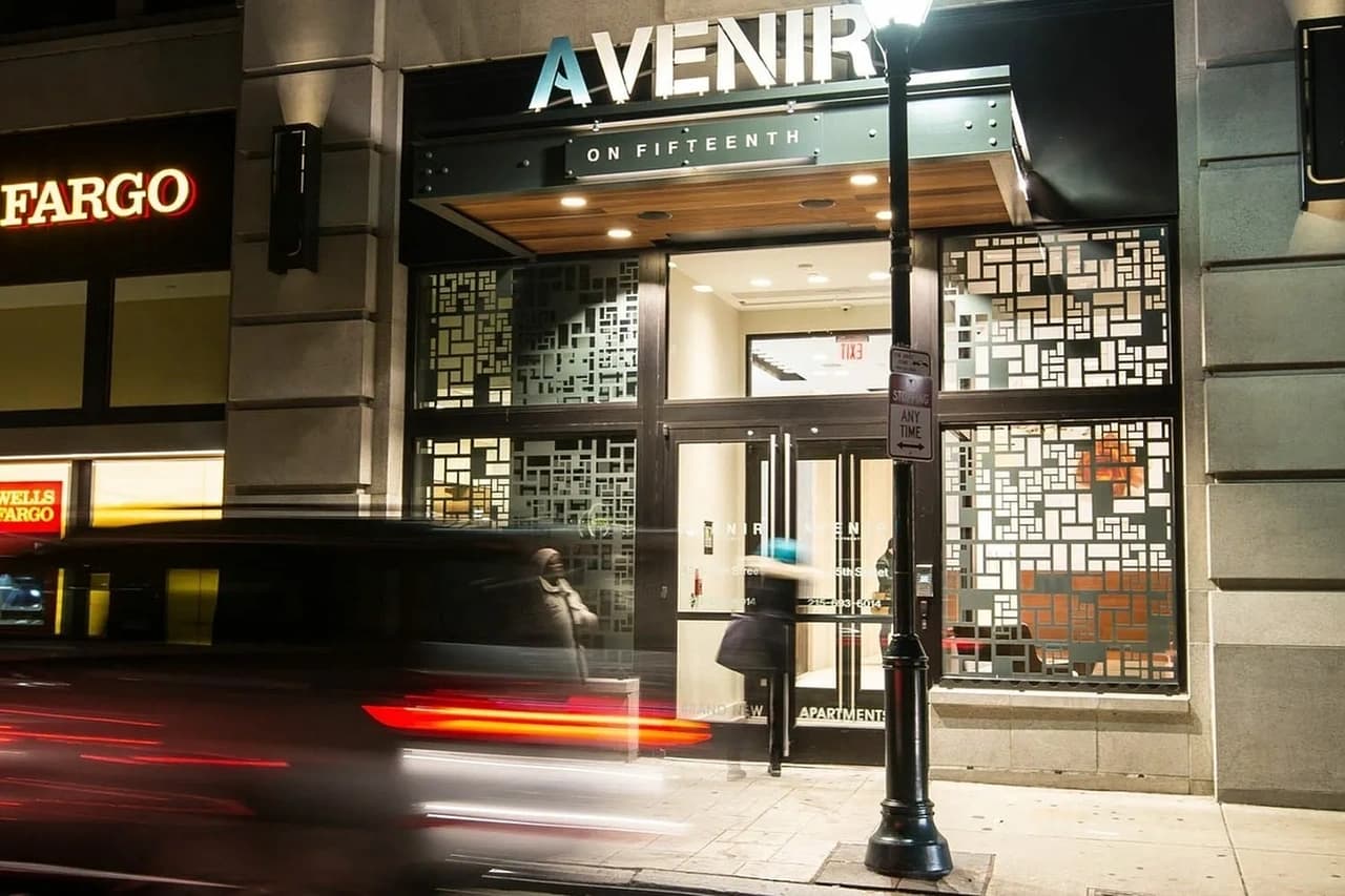Modern apartment building entrance with geometric patterned glass doors, illuminated signage, and urban street frontage.
