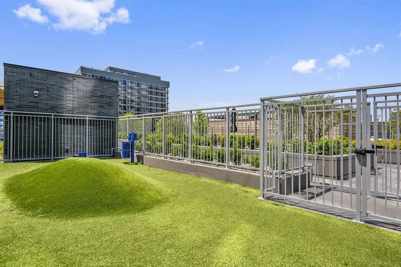 Rooftop terrace with bright green artificial turf, metal railing, pergola structure, and multi-story residential building in background under blue sky.