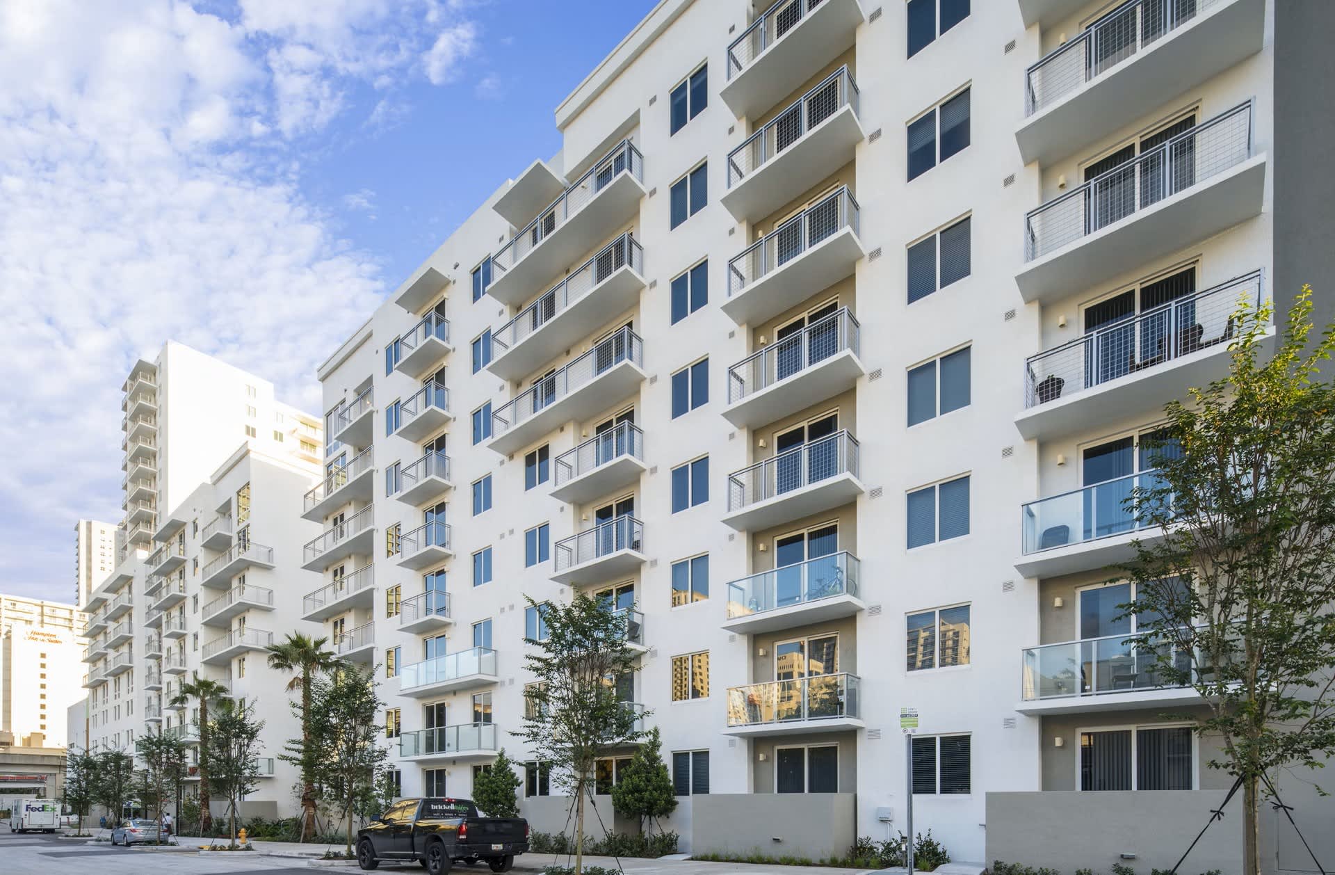 White multi-story apartment building with stacked balconies, large windows, and young trees along the street in Miami.