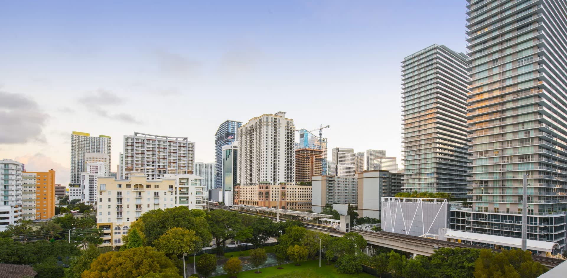 Elevated view of Miami's Brickell neighborhood with multiple high-rise residential towers, green trees, and downtown skyline under clear sky.