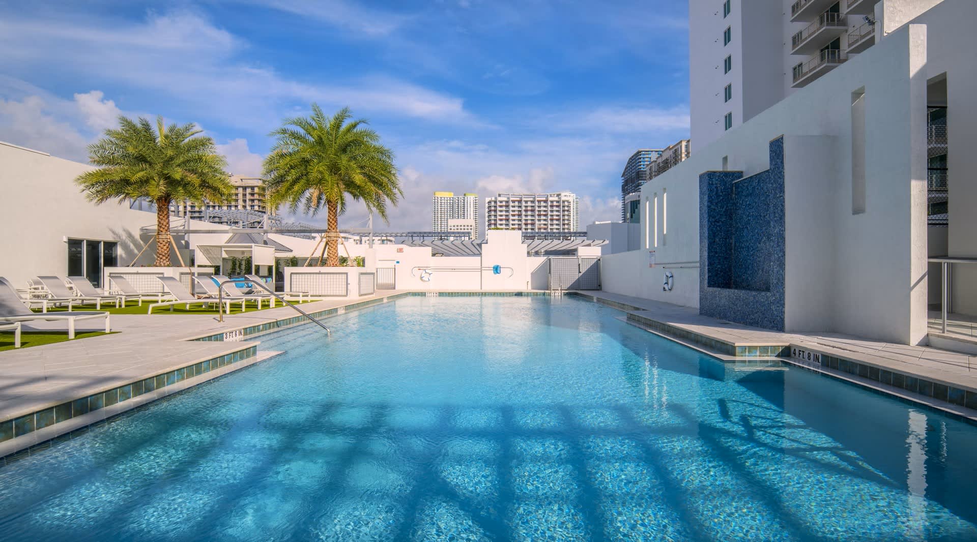 Resort-style swimming pool with clear blue water, white lounge chairs, palm trees, and modern white building with blue accent wall in urban setting.