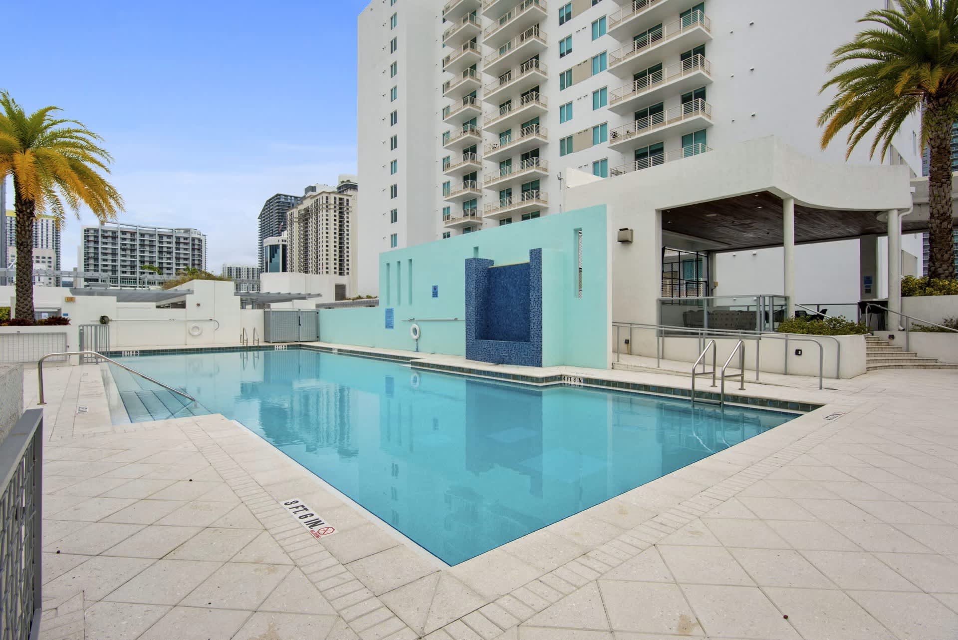 Rectangular swimming pool with clear turquoise water, light blue pool house structure, palm trees, and high-rise residential building in background.