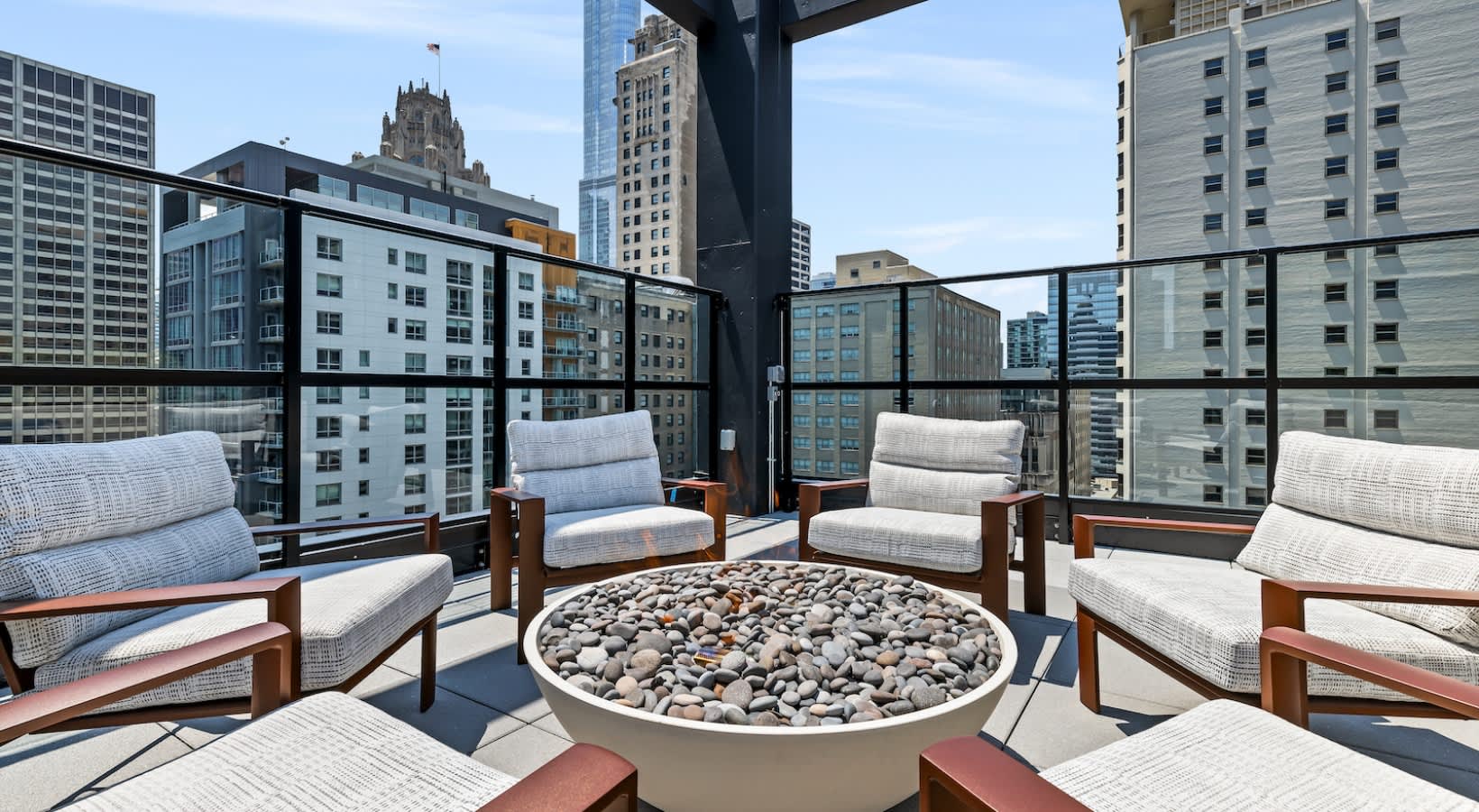 Rooftop terrace with wooden-frame lounge chairs, round fire pit with stones, and downtown Chicago skyline backdrop.