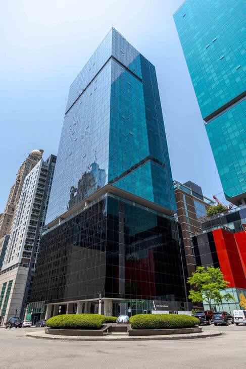 Contemporary high-rise building with turquoise glass facade, black metal framework, and ground-level plaza with manicured shrubs.