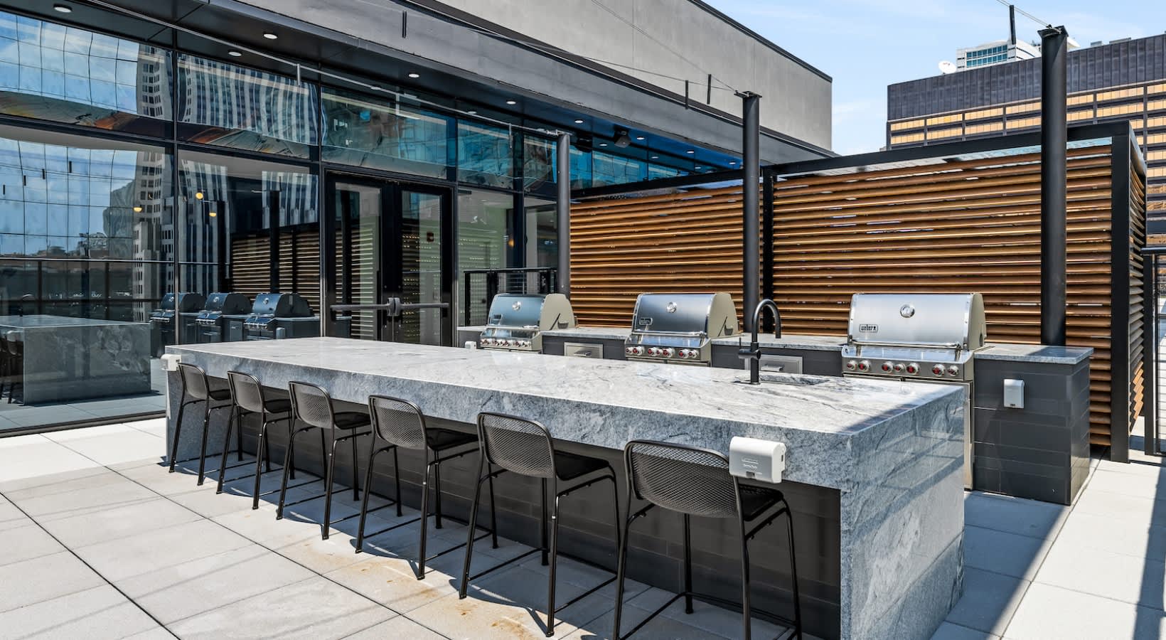Rooftop terrace with long marble bar, black bar stools, stainless steel grills, wooden privacy screen, and downtown Chicago skyline views.