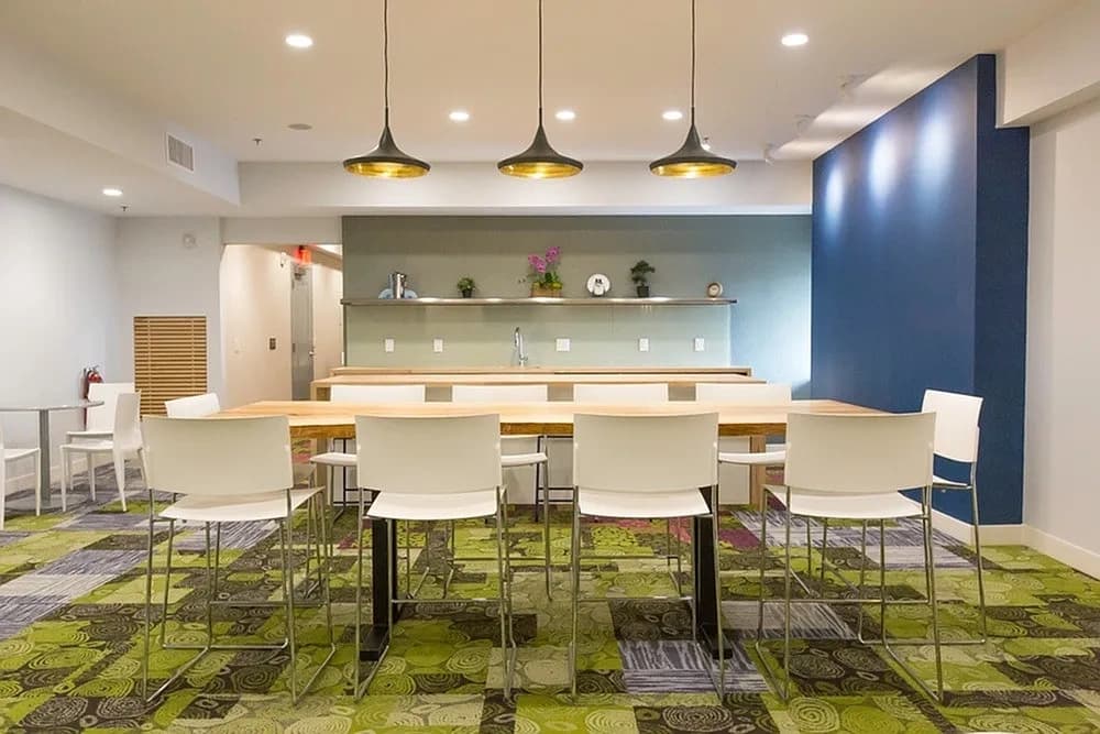 Common dining area with wooden table, white chairs, three pendant lights, sage green wall with shelving, and patterned green carpet.