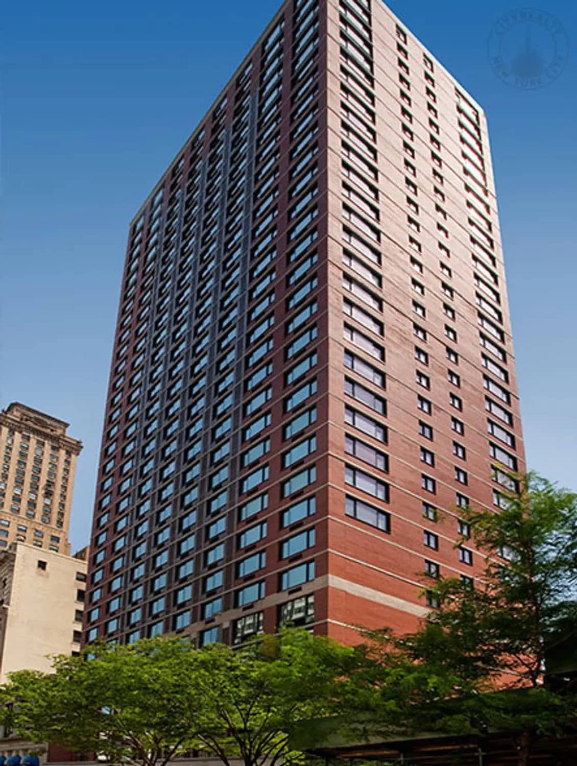 Tall brick residential tower with rows of windows and horizontal bands, surrounded by green trees and neighboring buildings under clear blue sky.