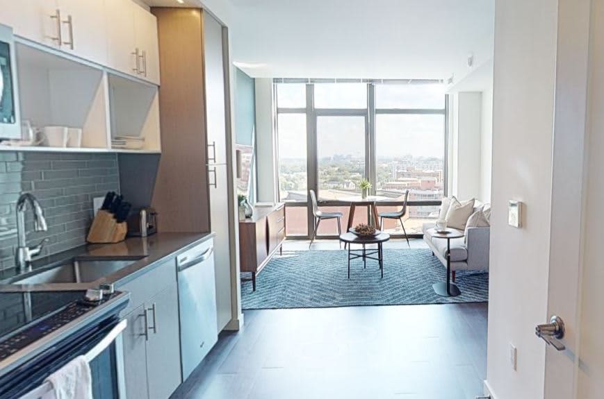 Modern kitchen with teal subway tile backsplash and white cabinetry opening to a living room with floor-to-ceiling windows and city views.