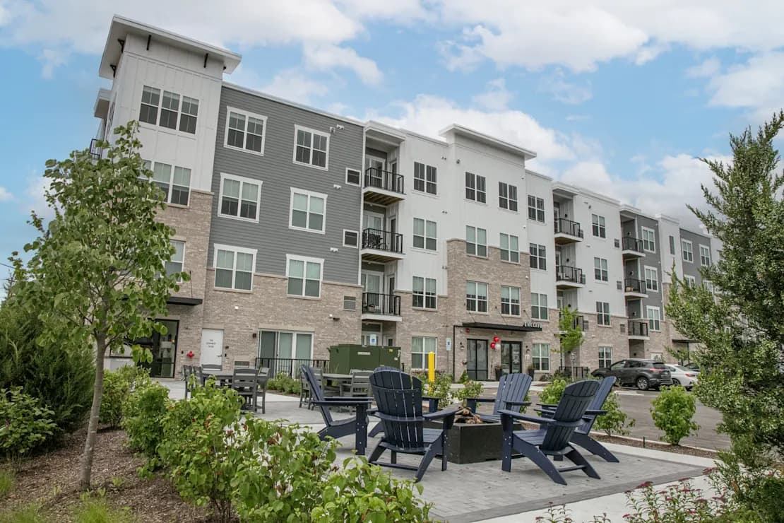 Modern apartment building exterior with courtyard featuring Adirondack chairs around a fire pit, surrounded by young trees and landscaping.