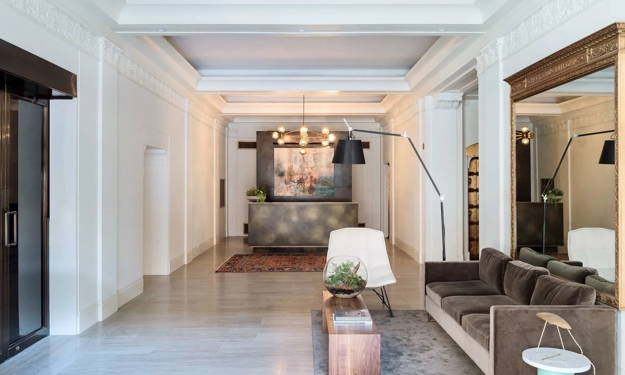 Modern apartment lobby with gray sofa, wooden coffee table, pendant lighting, framed artwork, and reception desk in background.