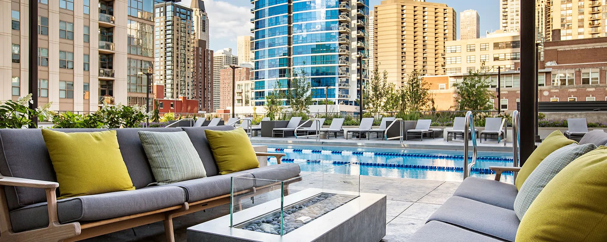 Rooftop pool deck with gray lounge seating, yellow accent pillows, fire pit, and lap pool overlooking Chicago skyline.