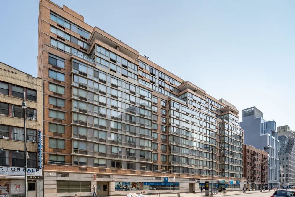 Multi-story residential building with red brick and glass facade, ground-floor retail spaces, and neighboring buildings along urban street.