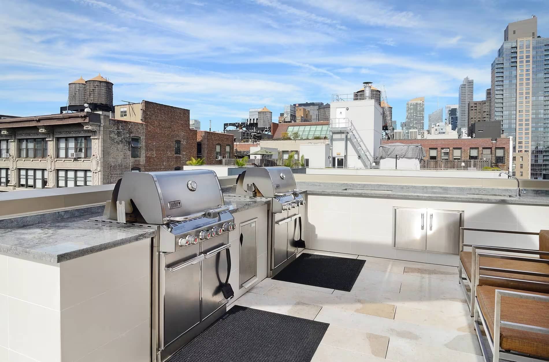 Rooftop kitchen with two stainless steel grills, white cabinetry, and expansive views of Manhattan buildings and skyline.