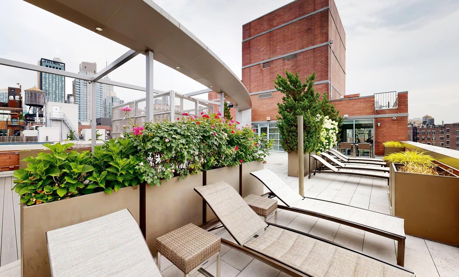 Rooftop terrace with lounge chairs, raised planter beds with pink flowers and greenery, pergola, and Manhattan skyline views.
