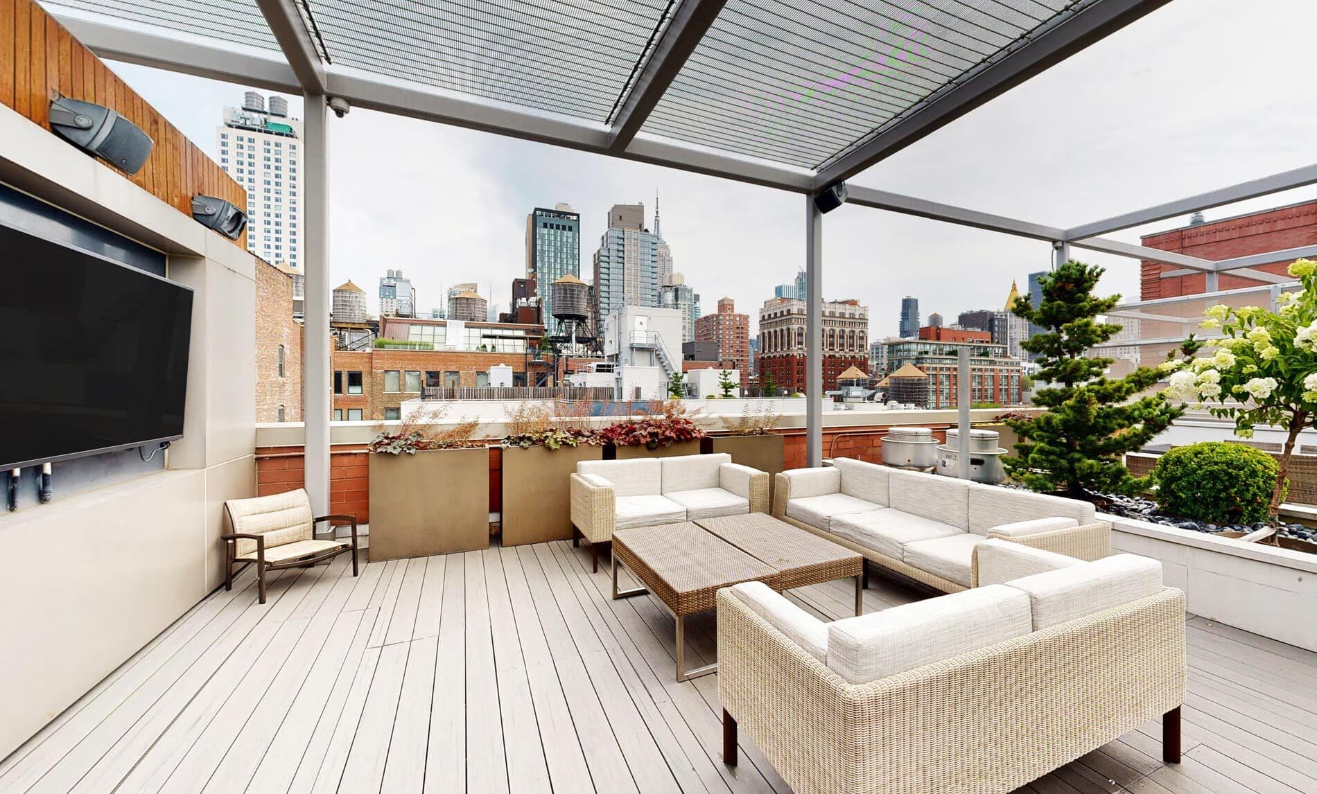 Rooftop terrace with woven lounge seating, wooden deck, retractable pergola, planters, and Manhattan skyline views.