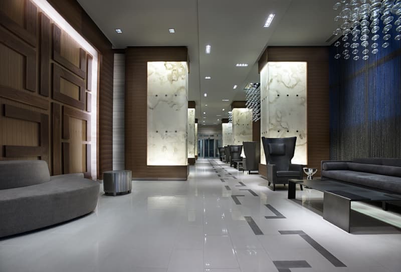 Modern lobby with white polished floors, dark wood wall panels, illuminated marble features, and contemporary gray seating.