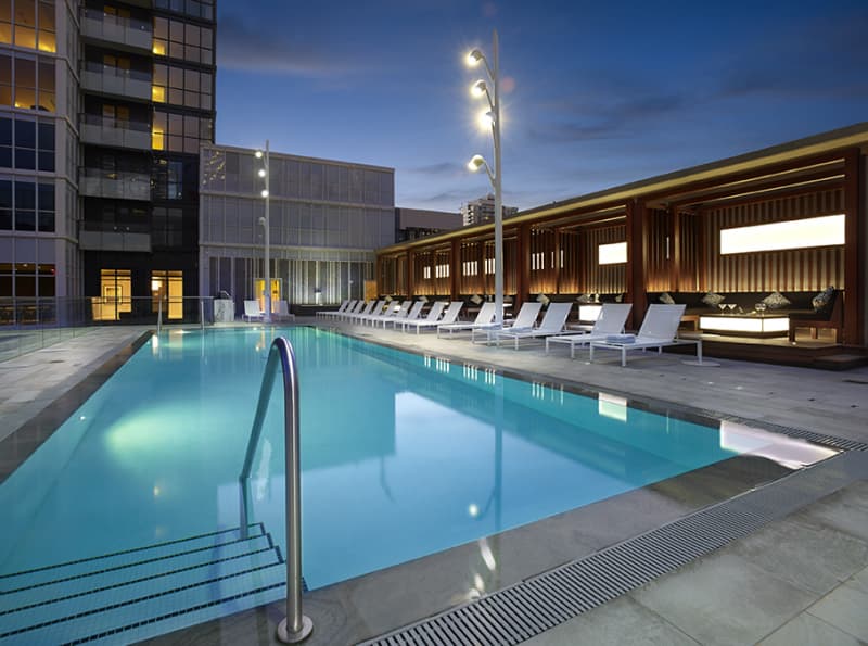 Illuminated rooftop pool with lounge chairs, metal railings, and modern building facades at dusk.