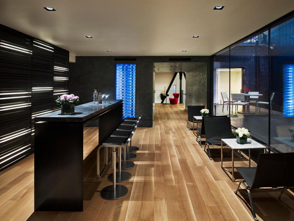 Contemporary lounge with black kitchen island, linear LED lighting, wood floors, and glass walls with city views.