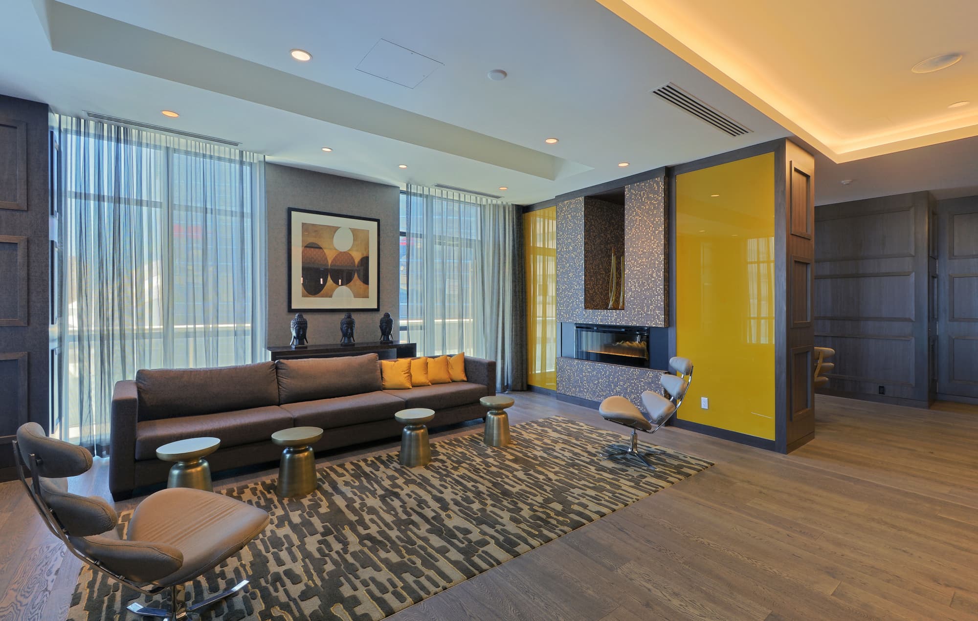 Modern living room with dark leather sofa, yellow accent wall, fireplace, floor-to-ceiling windows, and geometric patterned rug.