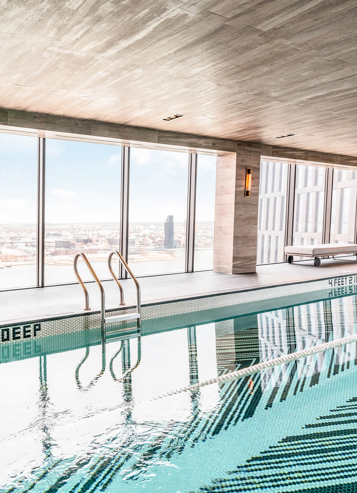 Indoor swimming pool with turquoise water, metal ladder, and floor-to-ceiling windows showing city skyline view.