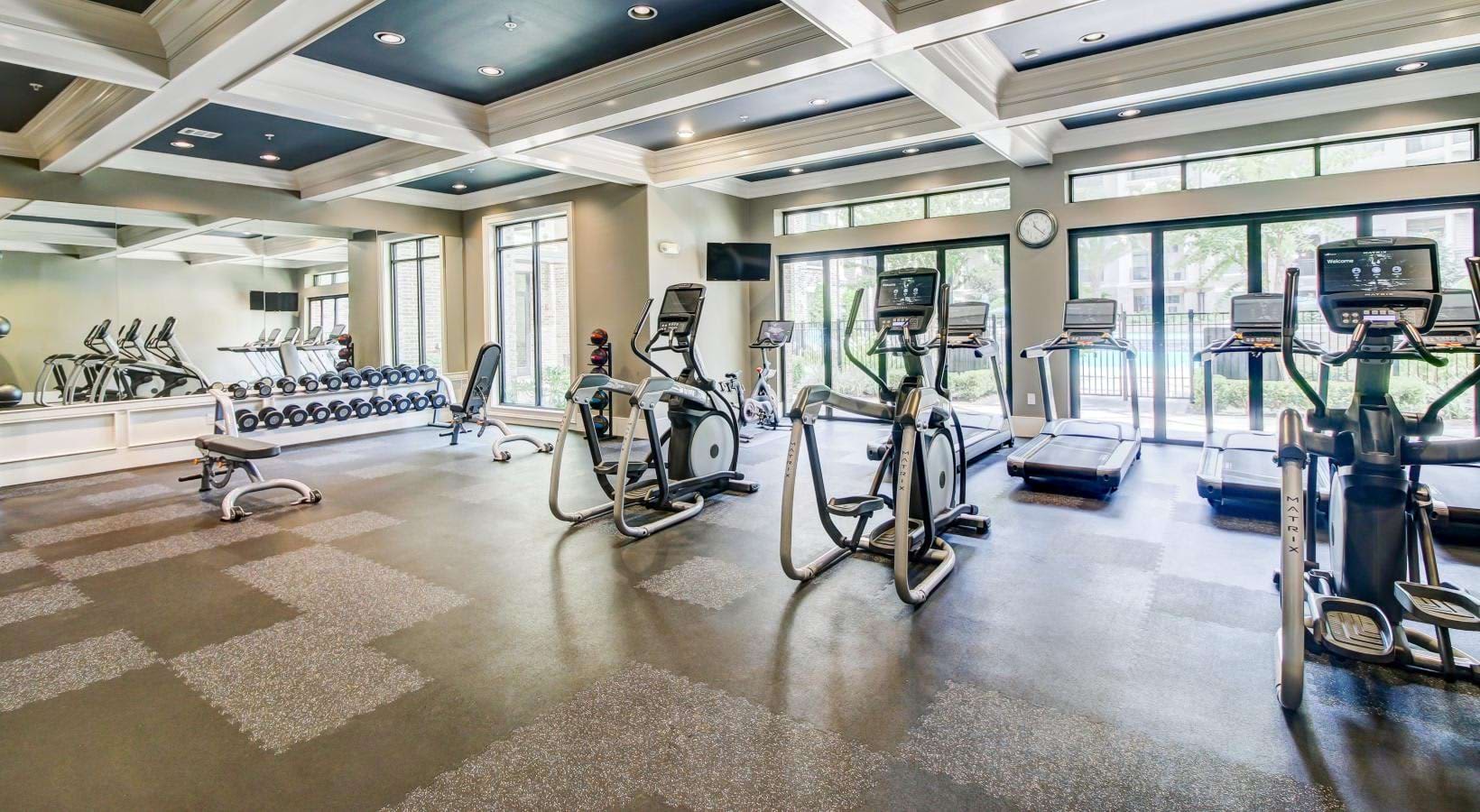 Fitness center with elliptical machines, stationary bikes, dumbbell rack, and coffered ceiling with large windows.