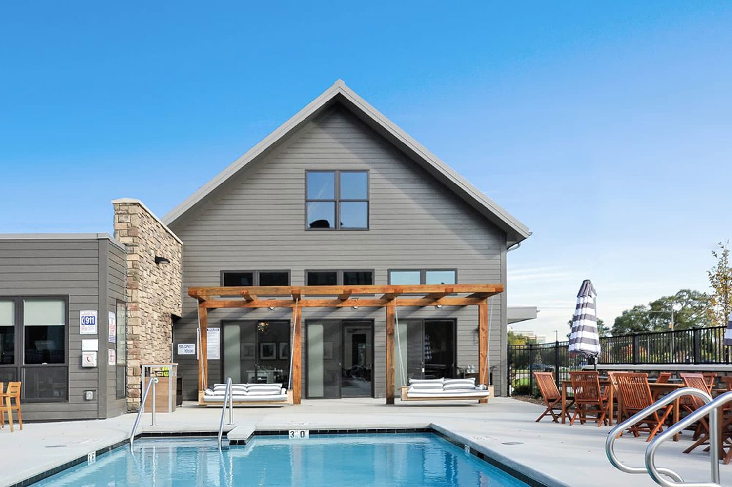 Modern gray house with wooden pergola, heated swimming pool in foreground, and lounge chairs on patio under clear blue sky.