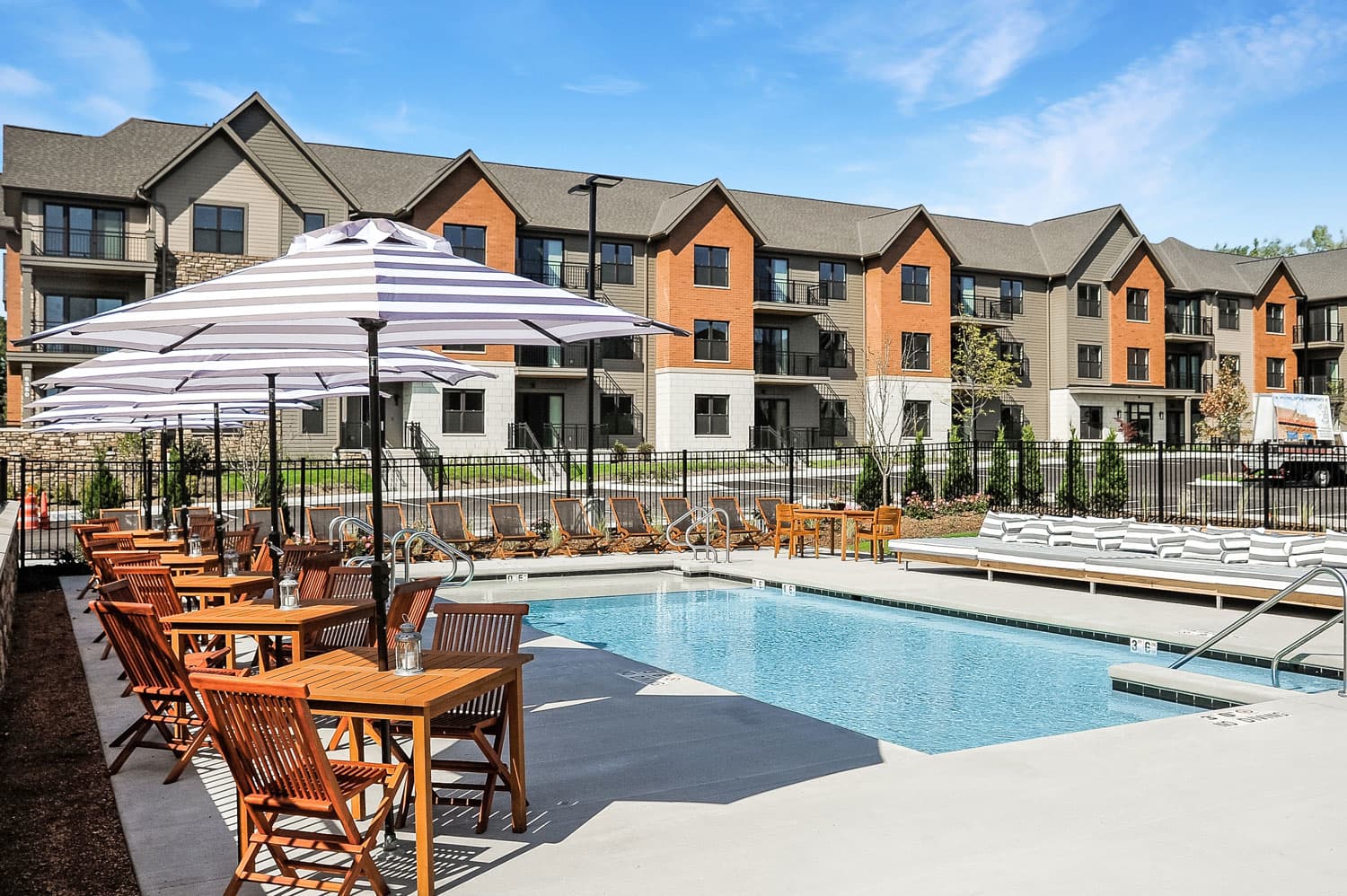 Resort-style swimming pool with wooden lounge chairs, striped umbrellas, and multi-story apartment buildings with orange and tan brick facades.