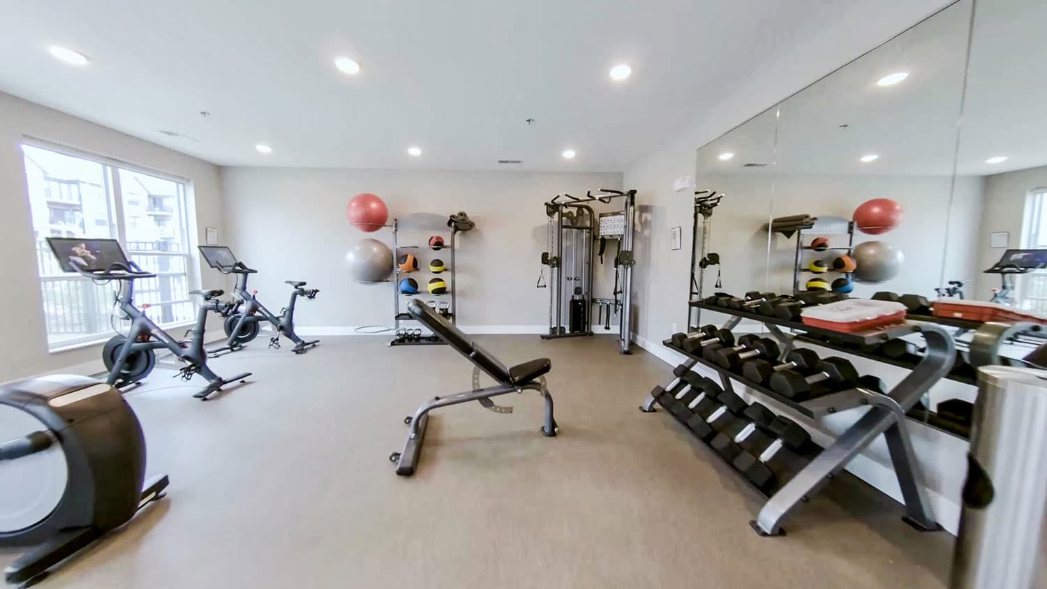 Bright fitness center with stationary bikes, weight rack, dumbbell set, exercise balls, and mirrored walls.