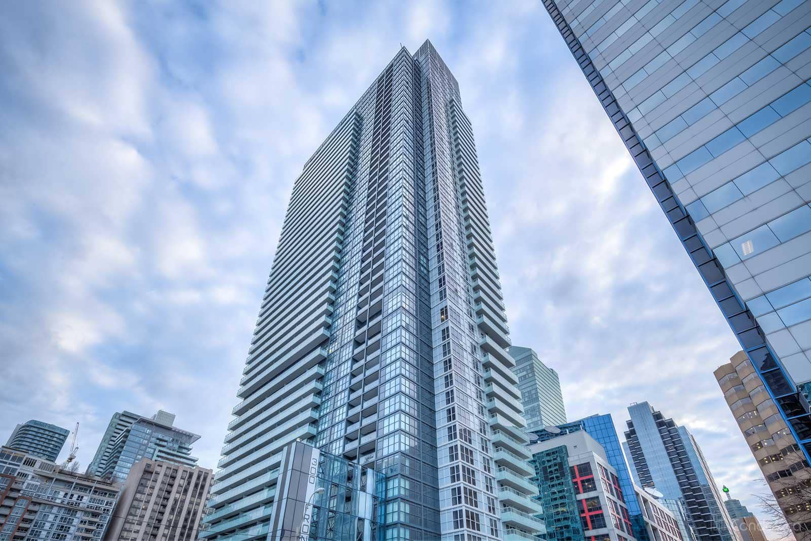 Tall residential high-rise tower with horizontal striping and glass balconies, surrounded by other downtown buildings under partly cloudy sky.