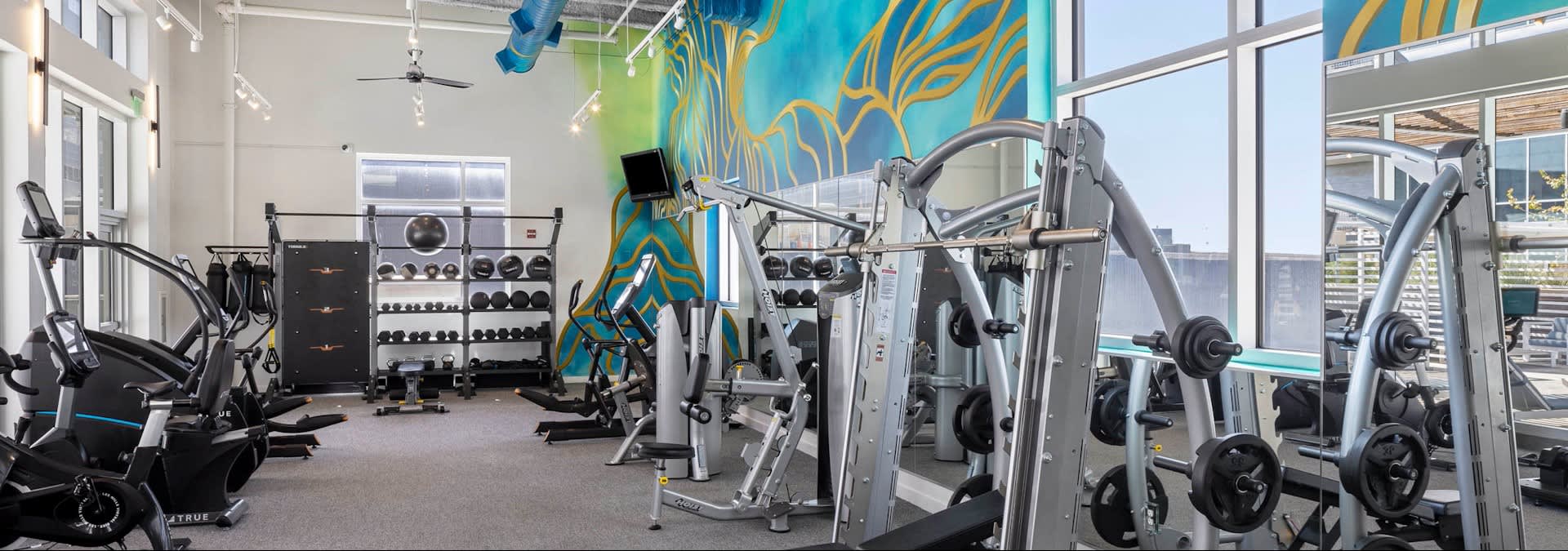 Fitness center with cardio machines, weight racks, and strength training equipment beneath blue and gold mural art and floor-to-ceiling windows.