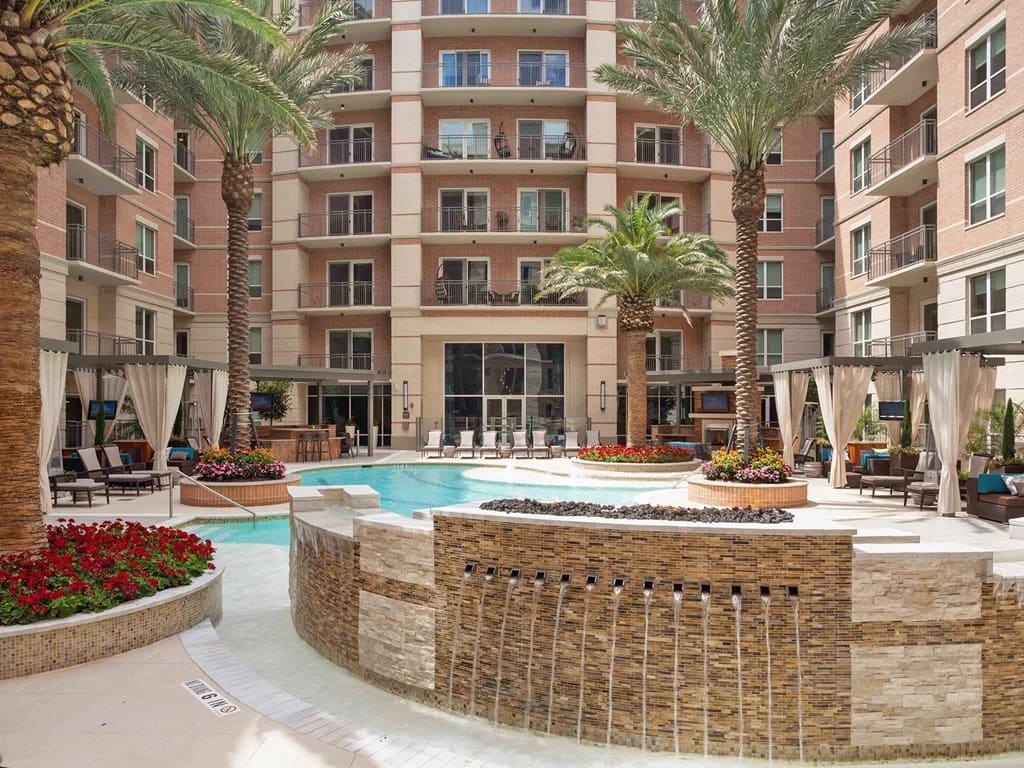 Courtyard pool area with brick bar counter, lounge chairs, white cabanas, red flowers, and palm trees surrounded by multi-story brick residential buildings.