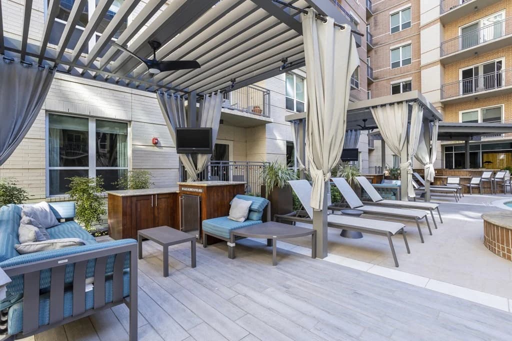 Outdoor courtyard with gray lounge chairs, blue cushioned seating, pergola with white curtains, and brick residential buildings in background.