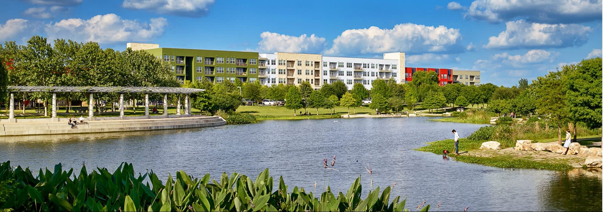 Waterfront view of AMLI at Mueller apartments with green and white buildings, lake, pergola, and landscaped grounds under blue sky.