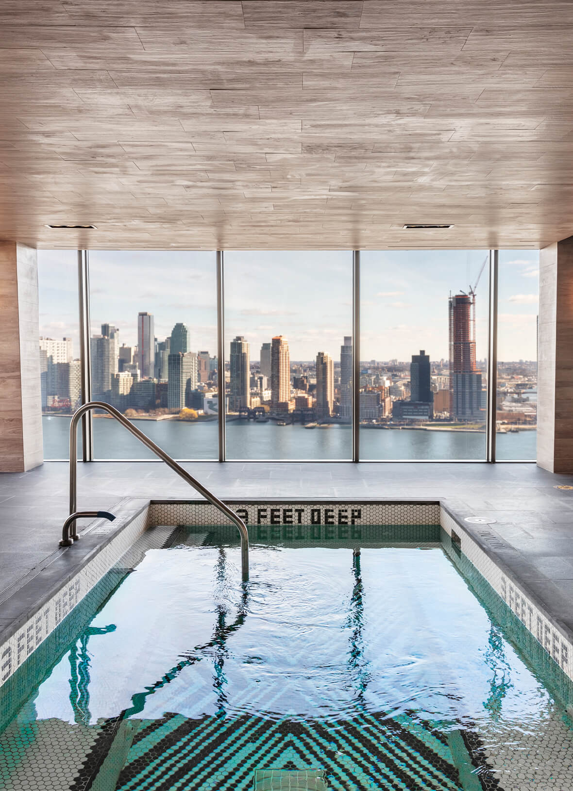 Indoor swimming pool with green tile trim and metal handrail, floor-to-ceiling windows overlooking Manhattan skyline and river.