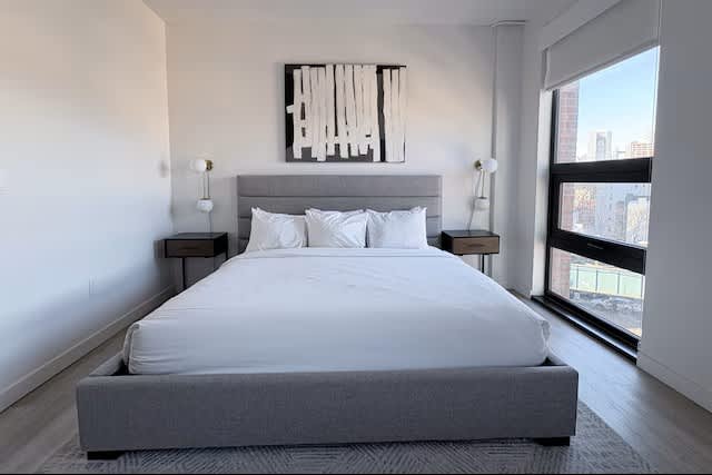 Bedroom with gray upholstered bed, white bedding, nightstands with pendant lamps, abstract wall art, and large windows overlooking city skyline.
