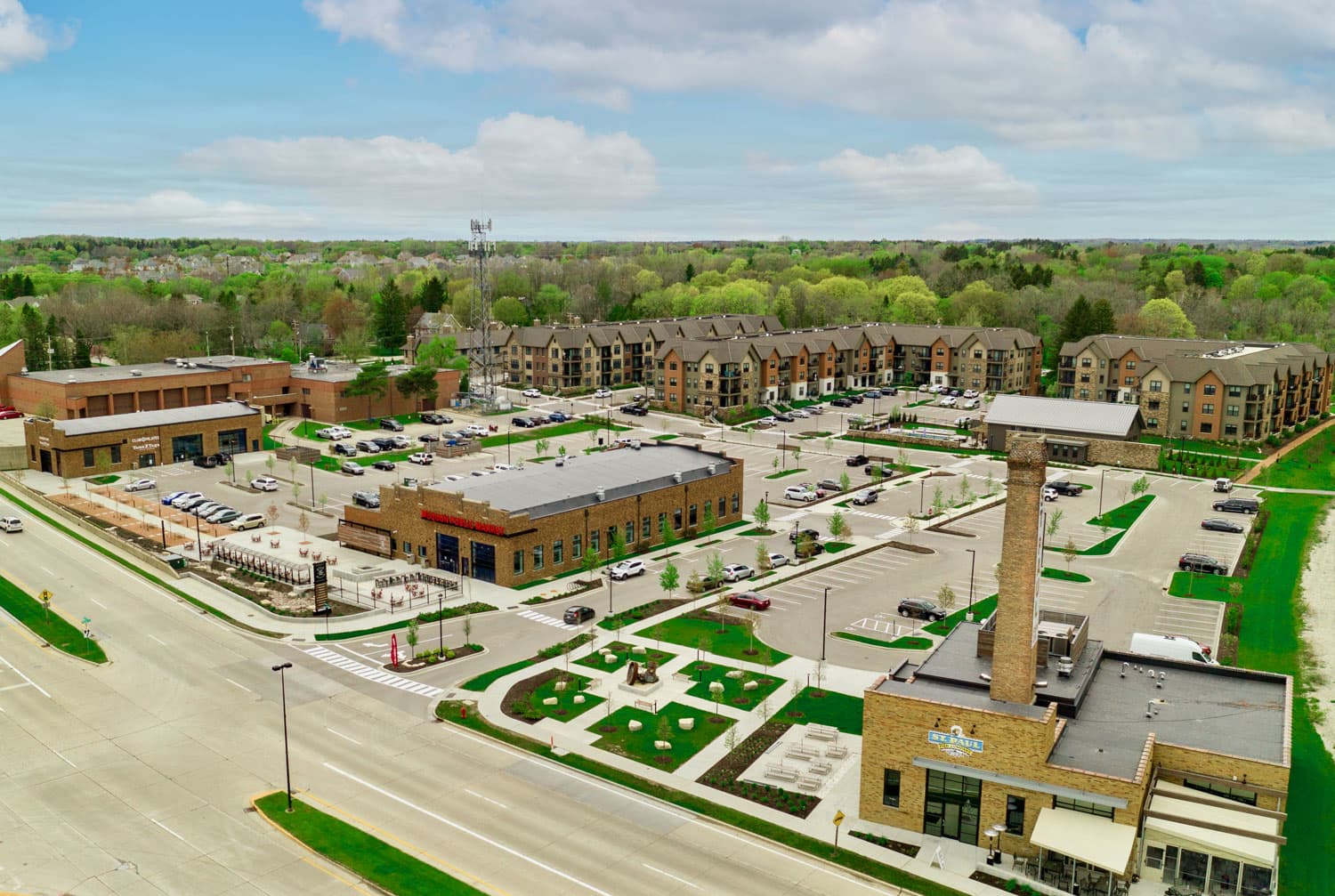 Aerial view of Spur 16 mixed-use development in Mequon with brick apartment buildings, parking areas, and green spaces surrounded by trees.