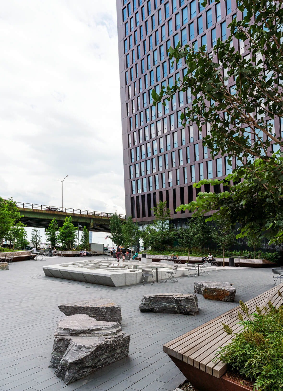 Outdoor plaza with paved ground, scattered rock seating, concrete water feature, green plants, and tall residential building overhead near highway bridge.